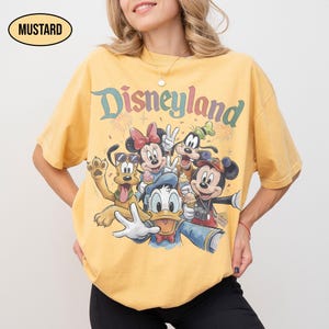 May include: Mustard-colored t-shirt featuring a graphic of Disney characters, including Mickey Mouse, Minnie Mouse, Donald Duck, Goofy, and Pluto. The word "Disneyland" is printed above the characters in colorful lettering. The shirt is a relaxed fit.