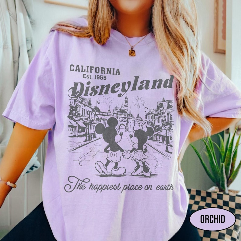 May include: A light purple t-shirt with a graphic of Mickey and Minnie Mouse holding hands in front of a Disneyland scene. The shirt has the text "CALIFORNIA Est. 1955 Disneyland" and "The happiest place on earth".