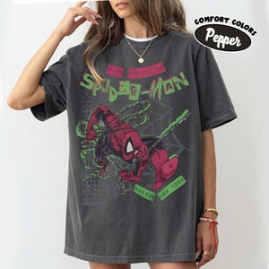 May include: Dark gray t-shirt featuring a graphic of Spider-Man in red and black, with green accents and the text "The Amazing Spider-Man." Additional text includes "Queens New York." The shirt has a vintage, distressed look.