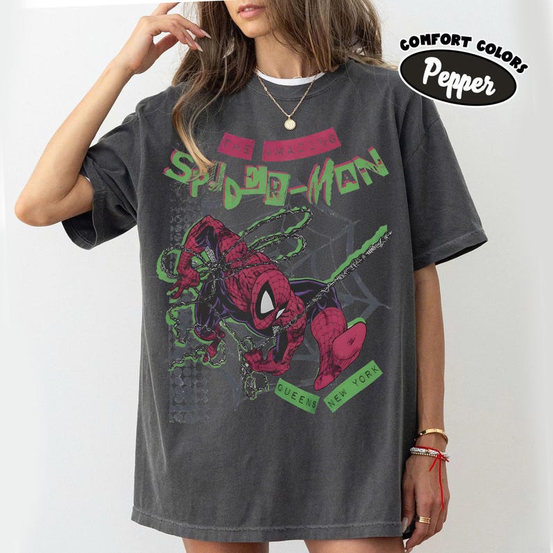 May include: Dark gray t-shirt featuring a graphic of Spider-Man in red and black, with green accents and the text "The Amazing Spider-Man." Additional text includes "Queens New York." The shirt has a vintage, distressed look.