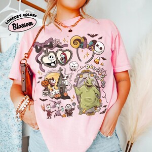 May include: Pink t-shirt with a graphic featuring characters and elements from "The Nightmare Before Christmas." The design includes Jack Skellington, Sally, Oogie Boogie, and other iconic imagery. The text "COMFORT COLORS Blossom" is printed on the top left.