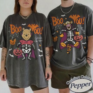 May include: Two charcoal gray t-shirts featuring Halloween graphics. The left shirt shows Winnie the Pooh in a skeleton costume and cape with the text Boo You. The right shirt shows Tigger in a skeleton costume and cape with the text Boo You.