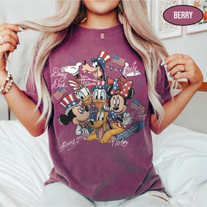 Może przedstawiać: Koszulka w kolorze jagodowym z grafiką postaci Disneya: Myszki Miki, Myszki Minnie, Kaczora Donalda, Kaczki Daisy, Goofy'ego i Pluto, świętująca 250. rocznicę USA. Projekt zawiera amerykańskie flagi i patriotyczne kapelusze.