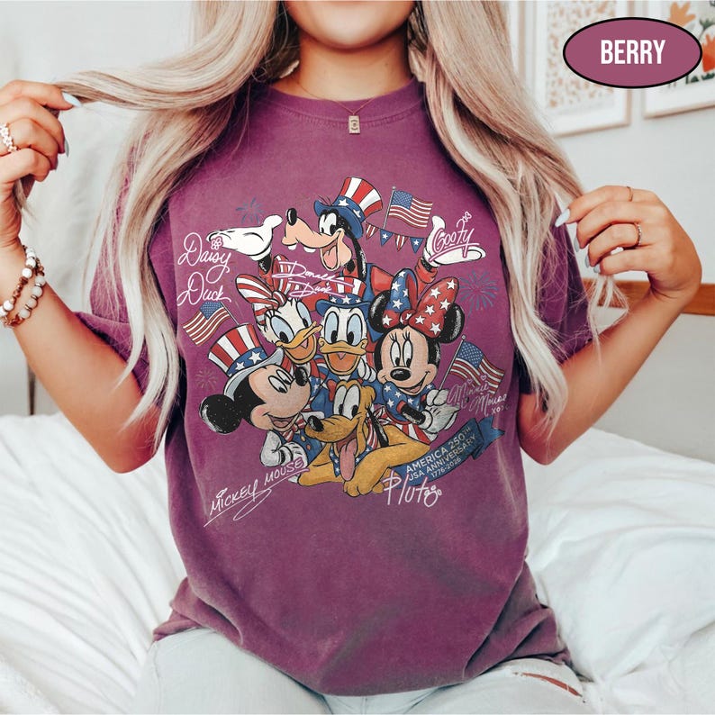 Może przedstawiać: Koszulka w kolorze jagodowym z grafiką postaci Disneya: Myszki Miki, Myszki Minnie, Kaczora Donalda, Kaczki Daisy, Goofy'ego i Pluto, świętująca 250. rocznicę USA. Projekt zawiera amerykańskie flagi i patriotyczne kapelusze.