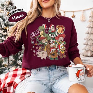 Op de afbeelding: Bordeaux sweatshirt met een feestelijke afbeelding van Toy Story-personages met kerstmutsen en cadeaus. De tekst "WOODY'S ROUDUP" is op de voorkant gedrukt. De sweatshirt heeft een ronde hals en lange mouwen. Het woord "SWEATSHIRT" en de kleur "Maroon" staan linksboven.