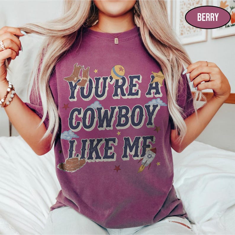 Puede incluir: Una camiseta de manga corta color baya con el texto "YOU'RE A COWBOY LIKE ME" en fuente azul oscuro. El gr&aacute;fico incluye ilustraciones de botas de vaquero, un sombrero, una pelota, una estrella de sheriff, nubes y un cohete estilo vintage.