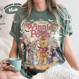 May include: A moss green t-shirt featuring a graphic of Winnie the Pooh characters dressed in Halloween costumes. The print includes Pooh, Piglet, Tigger, and Eeyore with pumpkins, bats, and spiderwebs. The text reads Disney Winnie the Pooh, Candy, and 100 Acre Wood.