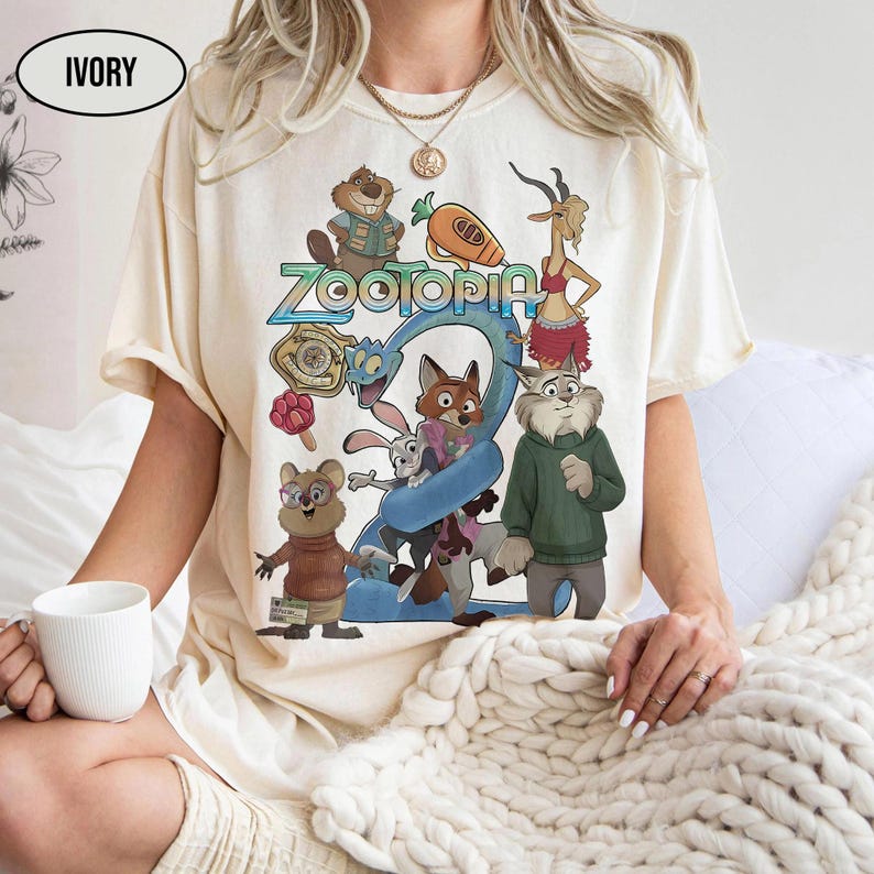 May include: Ivory-colored t-shirt featuring a colorful graphic of characters from the animated film "Zootopia." The design includes the film's title and various animal characters in a cartoon style. The shirt is short-sleeved.