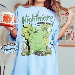 Puede incluir: Camiseta azul claro con un gr&aacute;fico de Oogie Boogie de "Pesadilla antes de Navidad" y el texto "Nightmare on Main Street". La camiseta tambi&eacute;n presenta una etiqueta Comfort Colors Chambray y una taza con un dise&ntilde;o de sol.