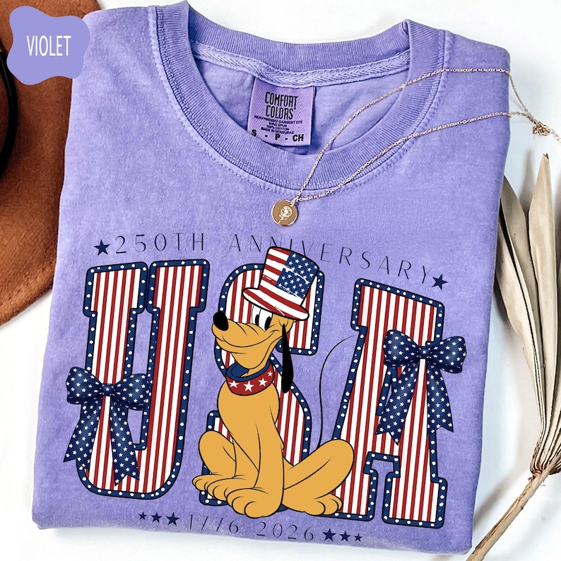 Może przedstawiać: Fioletowy t-shirt z kr&oacute;tkim rękawem z grafiką psa z kresk&oacute;wki w patriotycznym cylindrze i obroży. Wz&oacute;r zawiera napis 250TH ANNIVERSARY USA 1776 2026, duże litery w czerwono-białe paski oraz niebieskie kokardy w kropki.