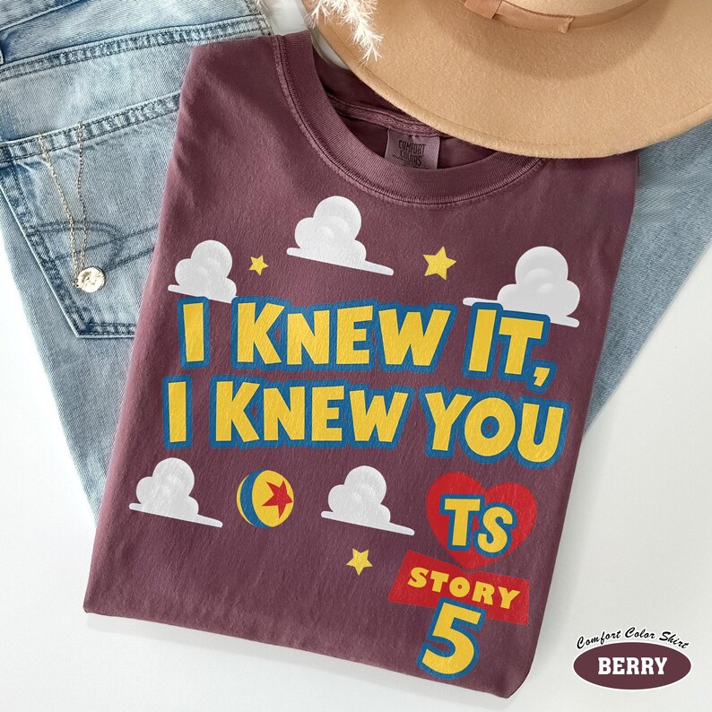 Puede incluir: Una camiseta color baya con el texto amarillo y azul I KNEW IT, I KNEW YOU, nubes blancas, estrellas amarillas, un coraz&oacute;n rojo, una pelota colorida y el texto TS STORY 5. La camiseta tiene cuello redondo y una etiqueta de la marca Comfort Colors.