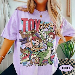 Puede incluir: Una camiseta lila con un gr&aacute;fico colorido de los personajes de Toy Story, incluyendo a Woody, Buzz Lightyear y Rex. Tambi&eacute;n se muestra el texto "TOY STORY" y "SINCE 1995". La camiseta tiene mangas cortas y cuello redondo.