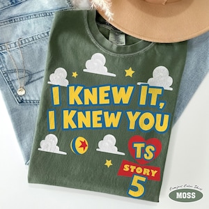 Puede incluir: Una camiseta verde oliva con el texto amarillo I KNEW IT, I KNEW YOU, gr&aacute;ficos de nubes blancas, estrellas amarillas, un coraz&oacute;n rojo con las letras TS, una pancarta roja con la palabra STORY, el n&uacute;mero 5 y una pelota amarilla con estrella roja.