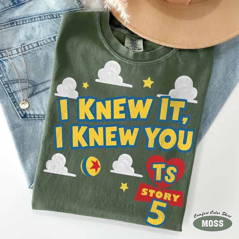Puede incluir: Una camiseta verde oliva con el texto amarillo I KNEW IT, I KNEW YOU, gr&aacute;ficos de nubes blancas, estrellas amarillas, un coraz&oacute;n rojo con las letras TS, una pancarta roja con la palabra STORY, el n&uacute;mero 5 y una pelota amarilla con estrella roja.