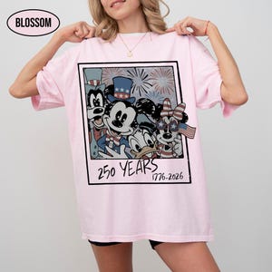 May include: A light pink t-shirt featuring a graphic print of cartoon characters wearing patriotic hats and star-spangled glasses. The design is framed like a polaroid with fireworks in the background and the text 250 YEARS 1776-2026 printed below.