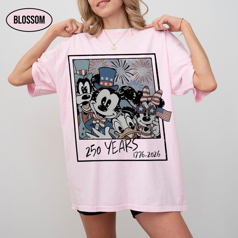 May include: A light pink t-shirt featuring a graphic print of cartoon characters wearing patriotic hats and star-spangled glasses. The design is framed like a polaroid with fireworks in the background and the text 250 YEARS 1776-2026 printed below.