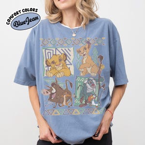 May include: A blue jean colored short-sleeve t-shirt featuring a graphic print of characters from The Lion King, including Simba, Nala, Pumbaa, Timon, Rafiki, and Zazu, arranged in four panels with geometric patterns and a Comfort Colors logo.