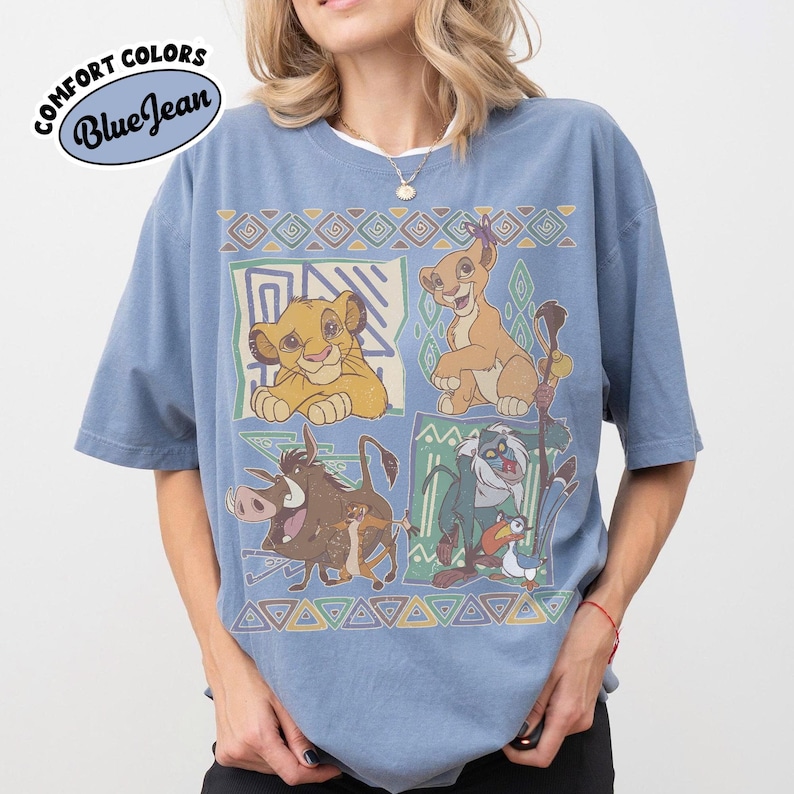 May include: A blue jean colored short-sleeve t-shirt featuring a graphic print of characters from The Lion King, including Simba, Nala, Pumbaa, Timon, Rafiki, and Zazu, arranged in four panels with geometric patterns and a Comfort Colors logo.