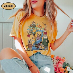 May include: Mustard-colored t-shirt featuring a colorful graphic of characters from the animated movie "Zootopia". The design includes the word "Zootopia" in a blue font, along with various animal characters. The shirt is a casual wear item.