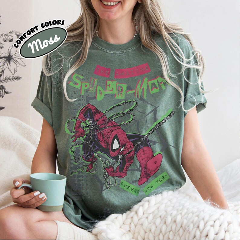 May include: Moss green t-shirt featuring a graphic of Spider-Man in a dynamic pose with chains and the text "Spider-Man" in a retro font. The shirt also includes the text "Queens New York". The shirt has rolled-up sleeves and a distressed look.