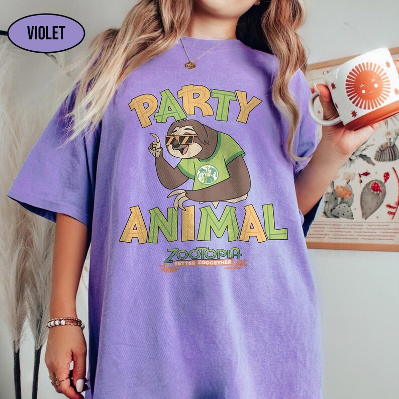 May include: A violet-colored t-shirt with the words "PARTY ANIMAL" in yellow and green lettering. The shirt features a cartoon sloth wearing sunglasses and a green shirt. Below the sloth is the text "ZOOTOPIA BETTER TOGETHER".
