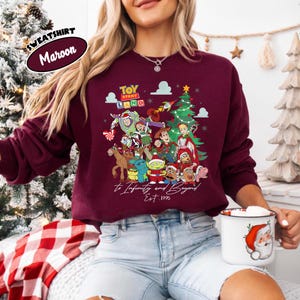May include: Maroon sweatshirt featuring a colorful graphic of Toy Story characters, a Christmas tree, and the text "Toy Story Land" and "to Infinity and Beyond Est. 1995". The sweatshirt has a crew neck and long sleeves.