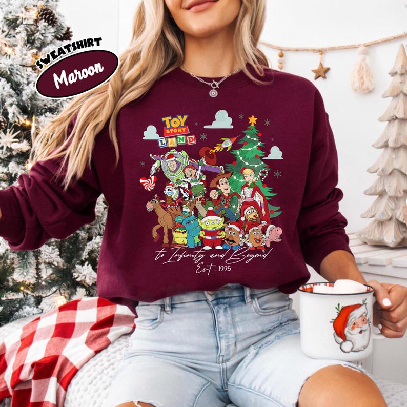May include: Maroon sweatshirt featuring a colorful graphic of Toy Story characters, a Christmas tree, and the text "Toy Story Land" and "to Infinity and Beyond Est. 1995". The sweatshirt has a crew neck and long sleeves.