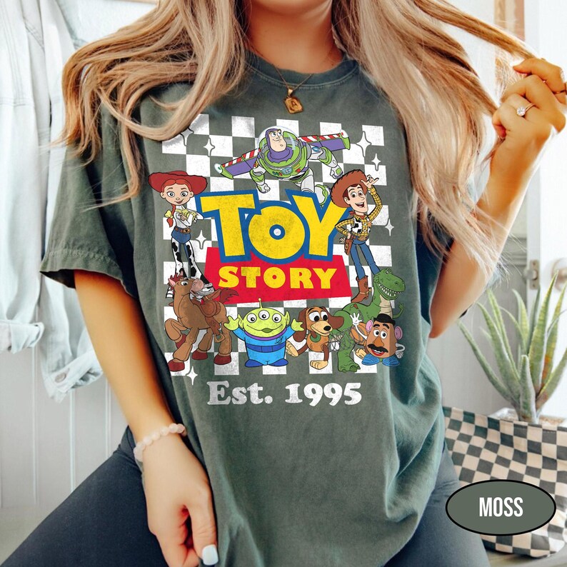 May include: Moss green t-shirt featuring the words "Toy Story" in yellow and blue, with cartoon characters from the movie. The shirt also has the text "Est. 1995" in white. The background is a checkered pattern.
