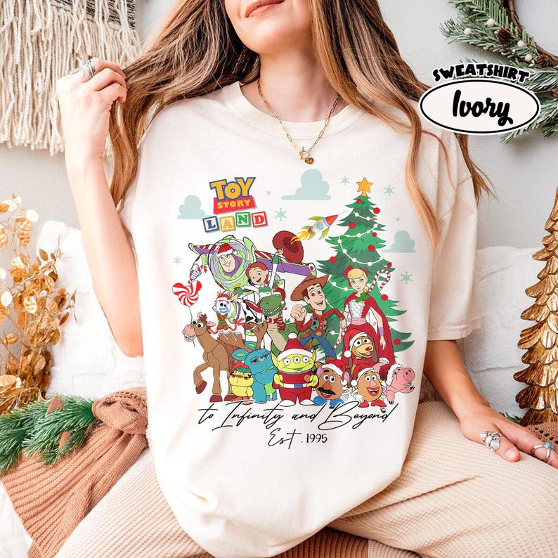 May include: Ivory-colored sweatshirt featuring a colorful graphic of Toy Story characters, a Christmas tree, and the text "Toy Story Land." The phrase "to Infinity and Beyond" and "Est. 1995" are also displayed. The word "Sweatshirt" and "Ivory" are in a white oval.