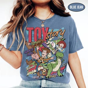 Puede incluir: Camiseta azul vaquero con un gr&aacute;fico de personajes de Toy Story. El dise&ntilde;o incluye a Woody, Buzz Lightyear, Jessie, Rex y otros personajes de la pel&iacute;cula. El texto "TOY STORY" se muestra de forma destacada, junto con "SINCE 1995".