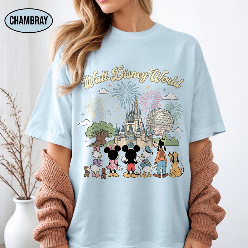 May include: Pale blue t-shirt featuring the words "Walt Disney World" and a graphic of Disney characters and fireworks. The graphic includes Mickey and Minnie Mouse, Donald Duck, Goofy, and other characters in front of a castle and a large sphere.