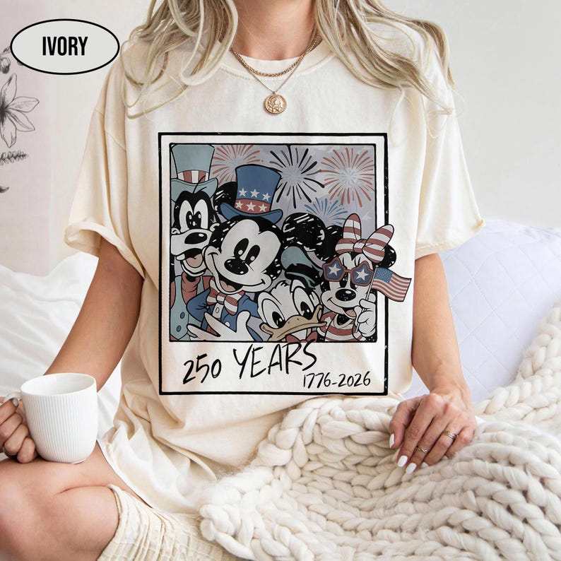 May include: An ivory short-sleeve t-shirt featuring a graphic print of cartoon characters in a polaroid frame. The design includes fireworks and the text 250 YEARS 1776-2026. The characters wear patriotic hats and accessories with red, white, and blue colors.