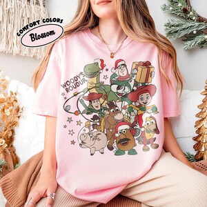 Op de afbeelding: Lichtroze T-shirt met een afbeelding van Toy Story-personages met kerstmutsen. Het ontwerp bevat de tekst "Woody's Roundup" en "Comfort Colors Blossom" in een cirkel.
