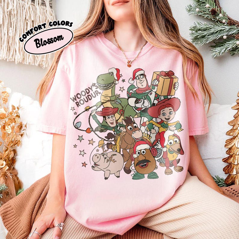 Op de afbeelding: Lichtroze T-shirt met een afbeelding van Toy Story-personages met kerstmutsen. Het ontwerp bevat de tekst "Woody's Roundup" en "Comfort Colors Blossom" in een cirkel.