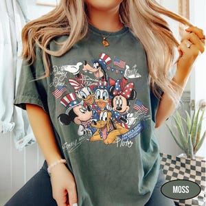 Może przedstawiać: Zielona koszulka w kolorze mchu z grafiką Myszki Miki, Myszki Minnie, Kaczora Donalda, Kaczki Daisy, Goofy'ego i Pluto w patriotycznych strojach. Projekt zawiera amerykańskie flagi i napis: "America 250th USA Anniversary 1776-2026".