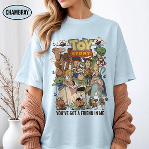 May include: Light blue t-shirt featuring a colorful graphic of Toy Story characters. The design includes the text "TOY STORY" and "YOU'VE GOT A FRIEND IN ME". The shirt has a crew neck and short sleeves.