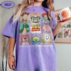 May include: A violet-colored t-shirt featuring a grid of Toy Story characters, including Woody, Buzz Lightyear, and others. The shirt has the text "TOY STORY" and "YOU'RE GOT A FRIEND IN ME" printed on it. A white mug with an orange sun design is held in the hand.