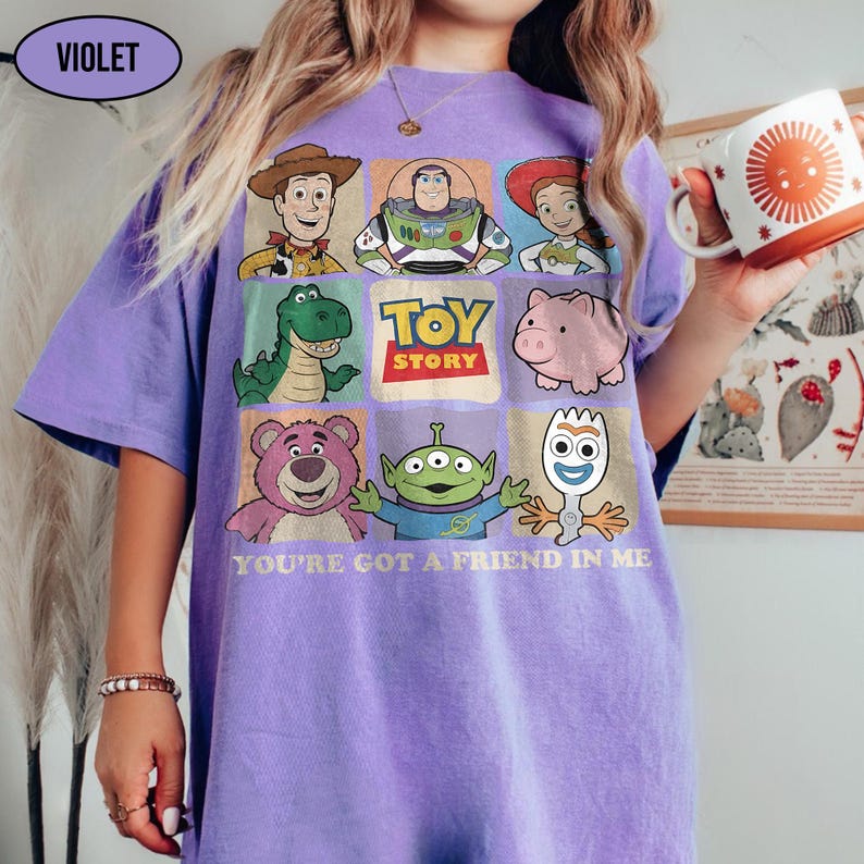 May include: A violet-colored t-shirt featuring a grid of Toy Story characters, including Woody, Buzz Lightyear, and others. The shirt has the text "TOY STORY" and "YOU'RE GOT A FRIEND IN ME" printed on it. A white mug with an orange sun design is held in the hand.