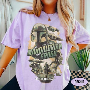 May include: A light purple t-shirt featuring a graphic from the Star Wars series, "The Mandalorian and Grogu." The design includes images of the Mandalorian, Grogu, and other characters, with the phrase "This is the Way." The word "ORCHID" is also visible.