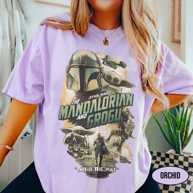 May include: A light purple t-shirt featuring a graphic from the Star Wars series, "The Mandalorian and Grogu." The design includes images of the Mandalorian, Grogu, and other characters, with the phrase "This is the Way." The word "ORCHID" is also visible.