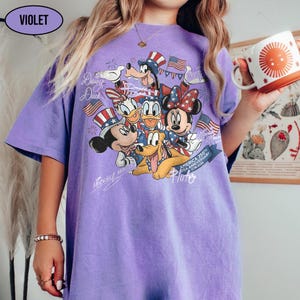 Może przedstawiać: Fioletowa koszulka z grafiką Myszki Miki, Myszki Minnie, Kaczora Donalda, Kaczki Daisy, Goofy'ego i Pluto w patriotycznych strojach. Koszulka ma napis "America 250th USA Anniversary 1776-2026".