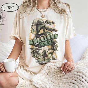 May include: Ivory-colored t-shirt featuring a Star Wars graphic with The Mandalorian and Grogu. The design includes images of the Mandalorian helmet, Grogu, and other characters, with the text "THIS IS THE WAY."