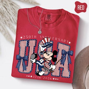 Może przedstawiać: Czerwony t-shirt z grafiką Myszki Miki w patriotycznym cylindrze i garniturze. Projekt zawiera napis 250TH ANNIVERSARY, USA oraz 1776-2026, otoczony dużymi literami w paski, niebieskimi kokardami w kropki oraz białymi gwiazdkami.
