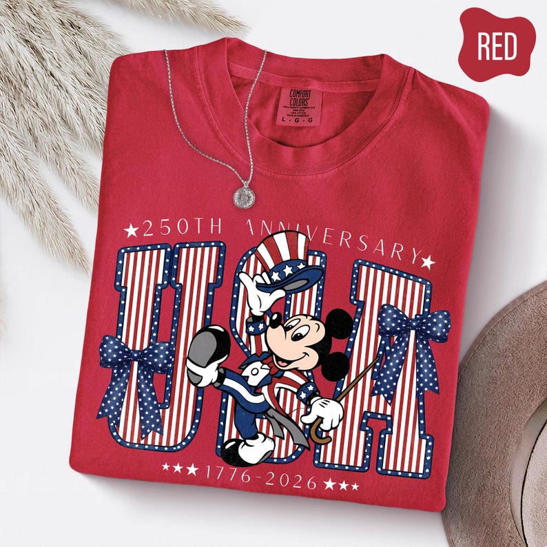 Może przedstawiać: Czerwony t-shirt z grafiką Myszki Miki w patriotycznym cylindrze i garniturze. Projekt zawiera napis 250TH ANNIVERSARY, USA oraz 1776-2026, otoczony dużymi literami w paski, niebieskimi kokardami w kropki oraz białymi gwiazdkami.