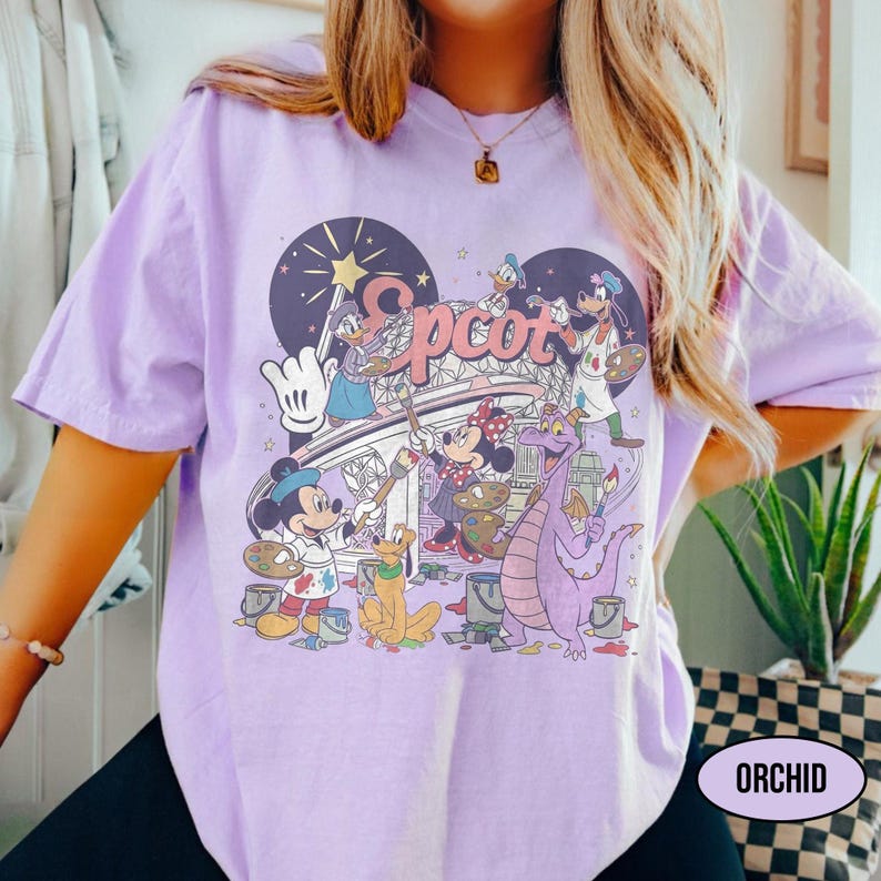 May include: A light purple t-shirt featuring a graphic of Disney characters painting the word "Epcot". Mickey Mouse, Minnie Mouse, Donald Duck, and Pluto are depicted. The shirt has a relaxed fit and the word "ORCHID" is printed on the bottom right.