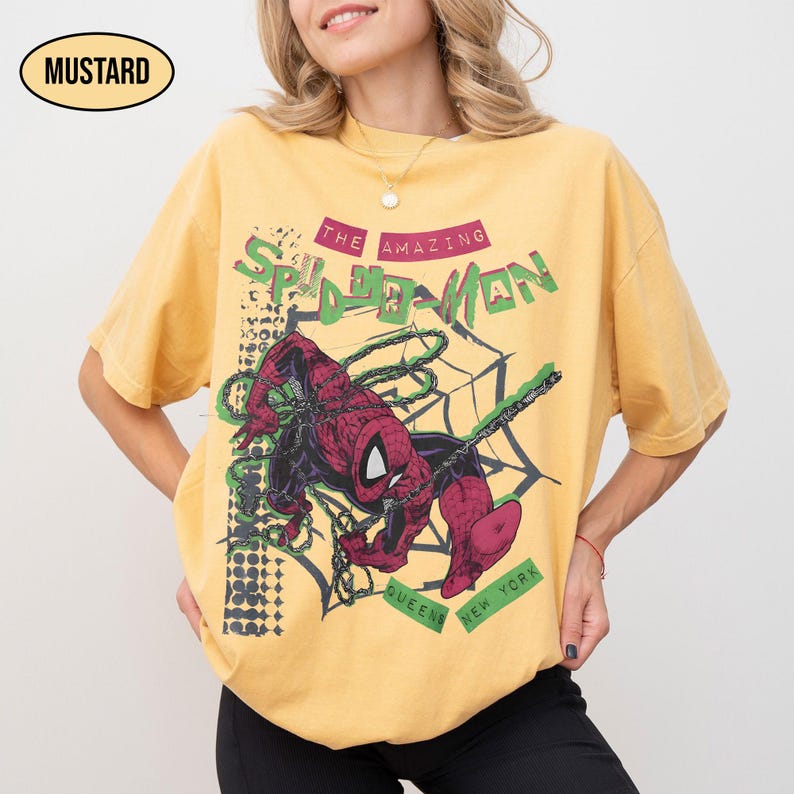 May include: Mustard-colored t-shirt featuring a graphic of Spider-Man in a web. The text "THE AMAZING SPIDER-MAN" is above the image, with "QUEENS NEW YORK" below. The shirt has a relaxed fit.