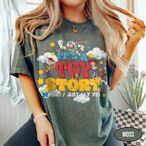 K&ouml;nnte beinhalten: Ein dunkelgr&uuml;nes T-Shirt mit einem Grafikdruck von Toy Story-Figuren, darunter Buzz Lightyear, Woody, Jessie und Slinky Dog. Der Text lautet IT'S A TOY STORY BABY JUST SAY YES, umgeben von gelben Sternen und wei&szlig;en Wolken.