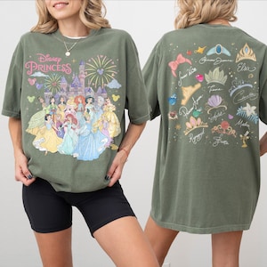 K&ouml;nnte beinhalten: Ein olivgr&uuml;nes T-Shirt mit einer Vorderseite, die Disney-Prinzessinnen vor einem Schloss mit Feuerwerk und dem Text Disney Princess zeigt. Die R&uuml;ckseite zeigt verschiedene Diademe und Kronen mit den Namen der jeweiligen Figuren darunter.