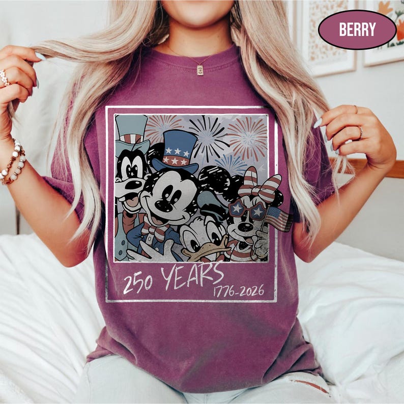 May include: A berry-colored t-shirt featuring a graphic print of cartoon characters wearing patriotic hats and glasses with fireworks in the background. The text 250 YEARS 1776-2026 is printed below the characters within a white rectangular frame.