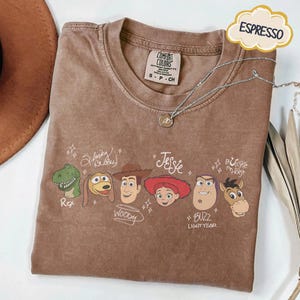 May include: An espresso-colored t-shirt featuring a graphic print of six animated characters with their names written in cursive script: Rex, Slinky Dog, Woody, Jessie, Buzz Lightyear, and Bullseye the Horse, accented with small star symbols.